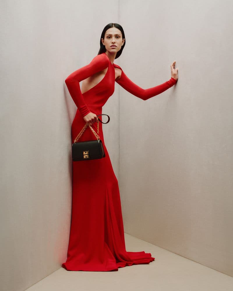 Elegant adult female fashion model in a long red dress strikes a pose with a black purse against a light-grey studio wall.