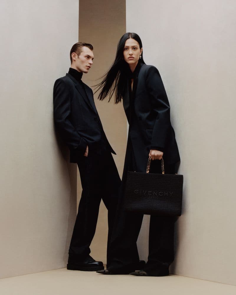 Fashion shot of a male and female model in black suits in a studio, with a black textured handbag. Fashion, studio lighting, posing.
