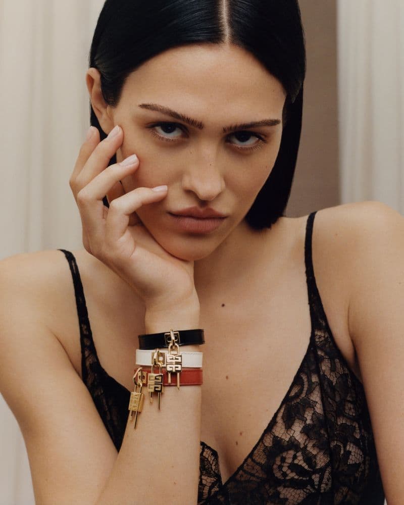 Close-up of a woman with dark hair, wearing a black lace top and bracelets, resting her face on her hand indoors.