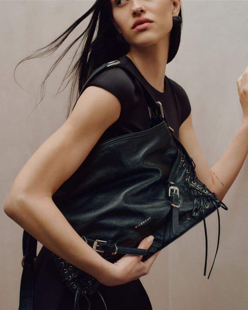 Fashion shot of a woman with long dark hair wearing a black outfit and carrying a black leather purse with metal buckles. 