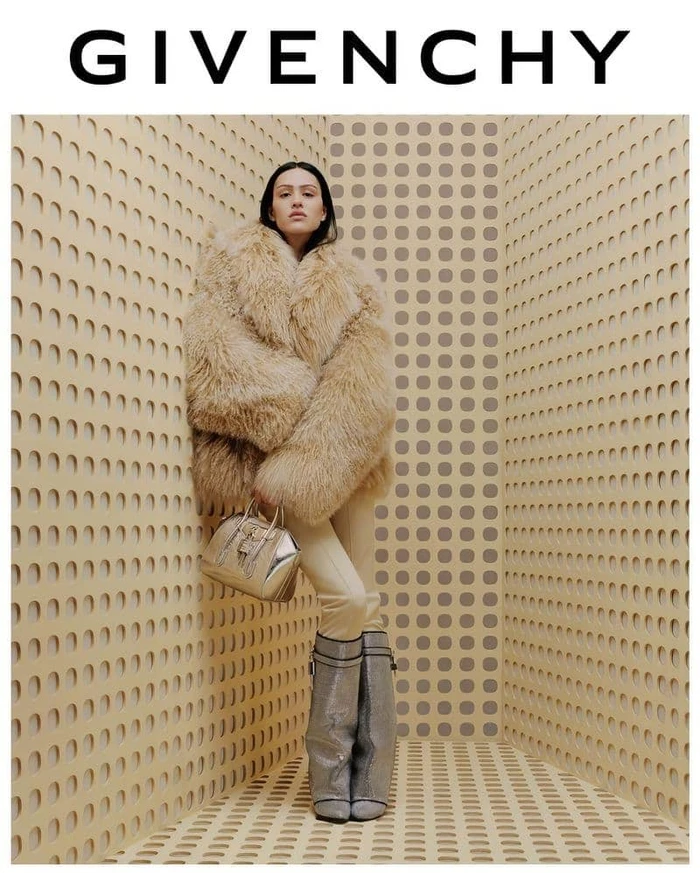 Model in a fluffy faux fur coat and metallic knee-high boots holding a handbag in a patterned corner setting.