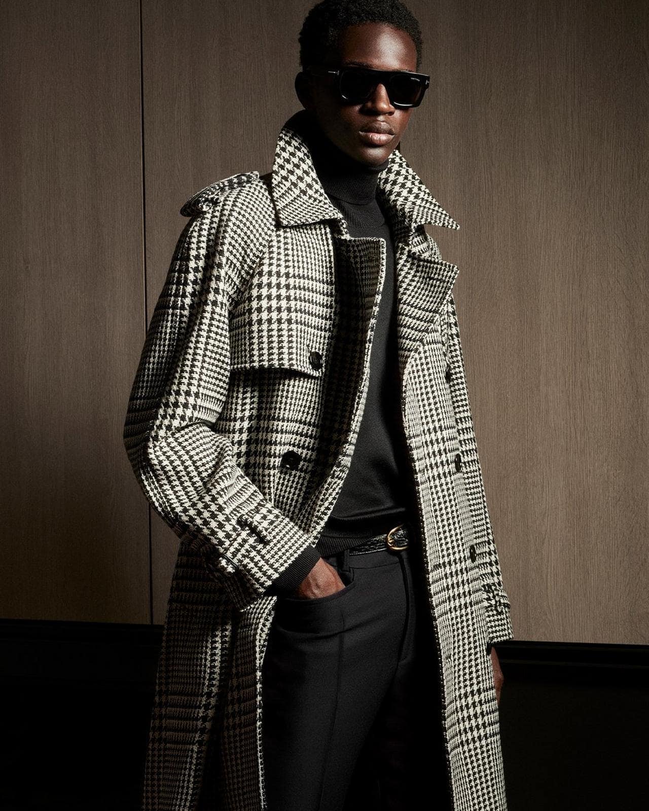 An African-American man poses indoors wearing a black turtleneck, black pants, and a long patterned overcoat and sunglasses.