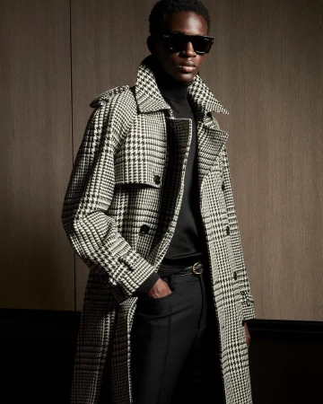 An African-American man poses indoors wearing a black turtleneck, black pants, and a long patterned overcoat and sunglasses.