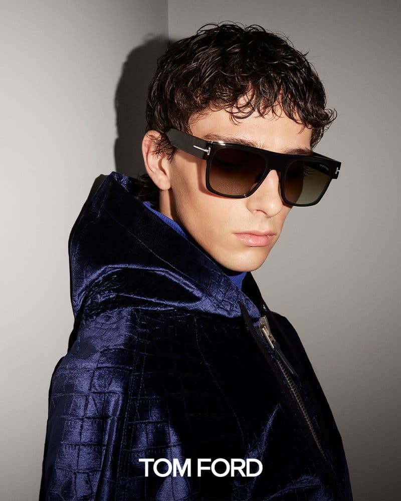 A fashionable young adult male wearing sunglasses and a stylish blue jacket. Focus on the model and clothing in a studio setting.