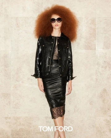 A woman with an afro hairstyle poses wearing a black leather jacket, skirt, and sunglasses, standing in front of a marble background.