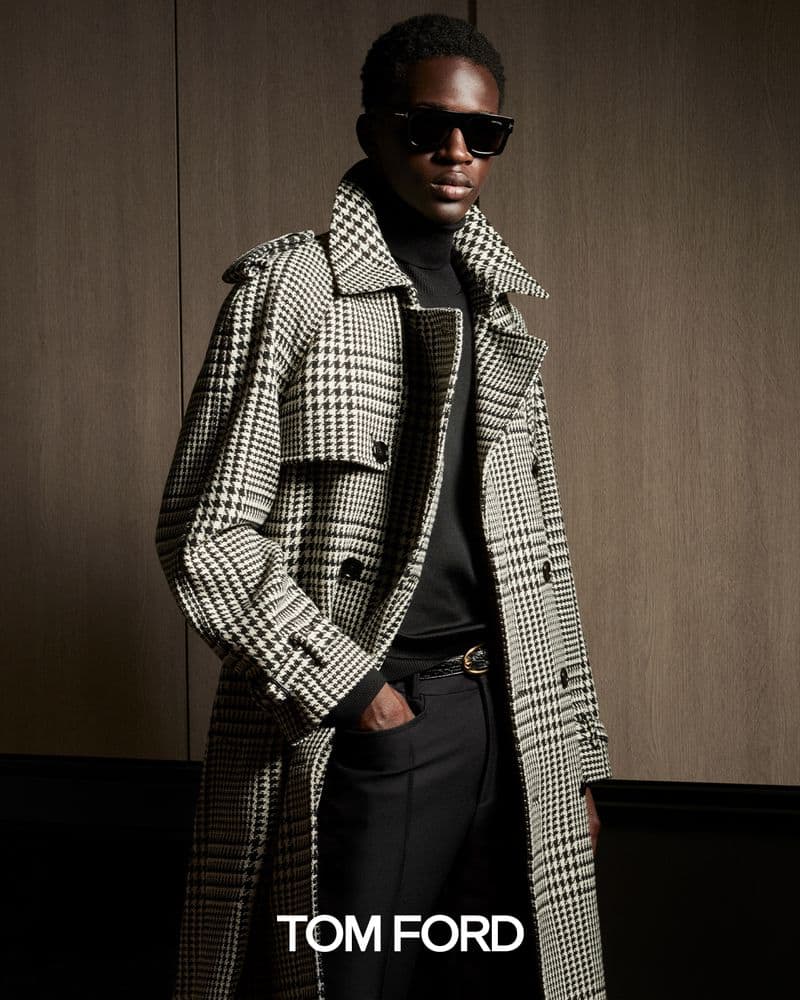 A stylish African-American male in a black turtleneck, plaid trench coat, and sunglasses strikes a pose against a brown backdrop for a fashion advertisement.