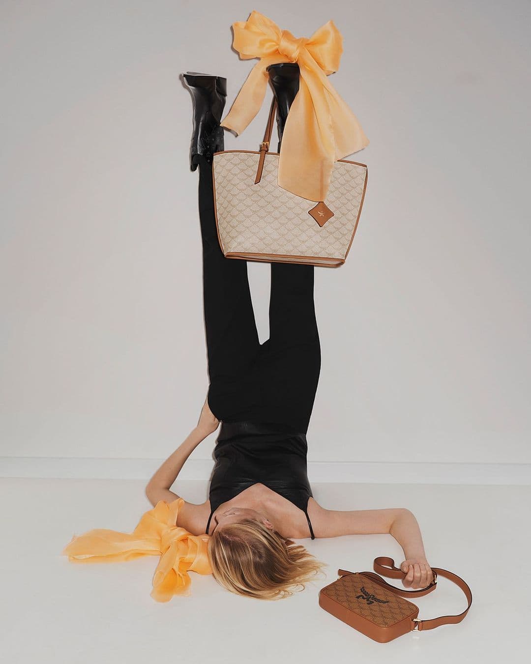 Fashion shot of woman lying on floor with handbag, wearing black leggings and top, shiny black boots, and orange scarf.