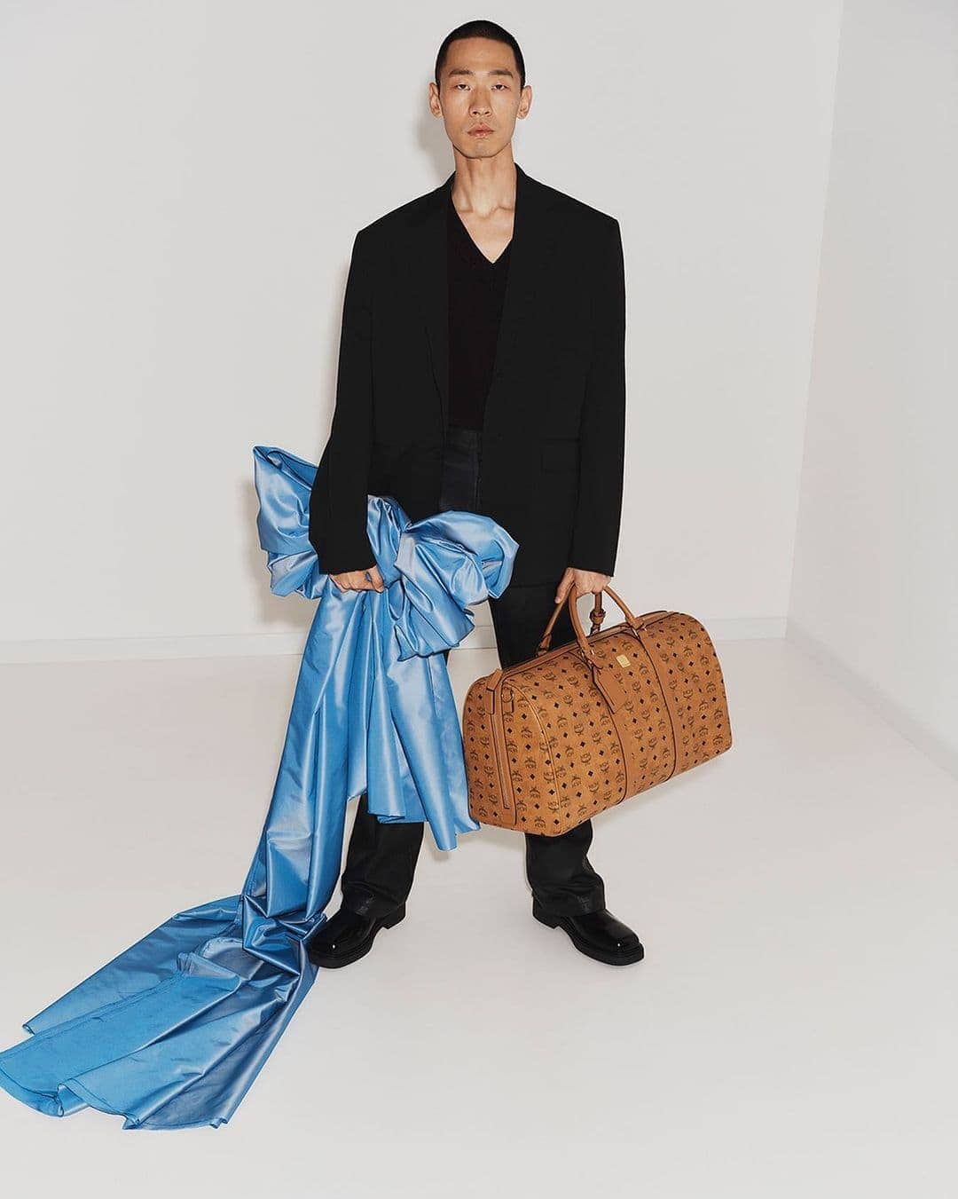 An Asian man in a black suit jacket and black pants holds a brown leather duffel bag and a large blue satin ribbon in a studio.