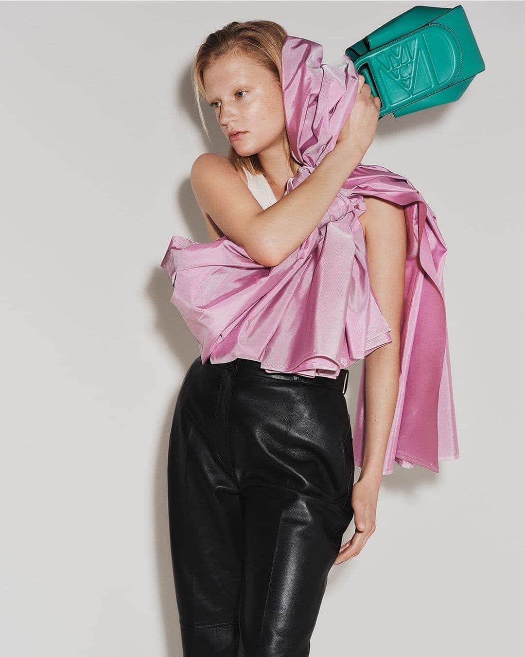 Fashionable woman poses in a pink top and black leather pants, holding a green purse against a solid white backdrop for a stylish studio shot.