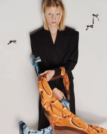 Fashion editorial shot of a blonde model in a black suit holding a patterned scarf in a studio with hair ribbon props.