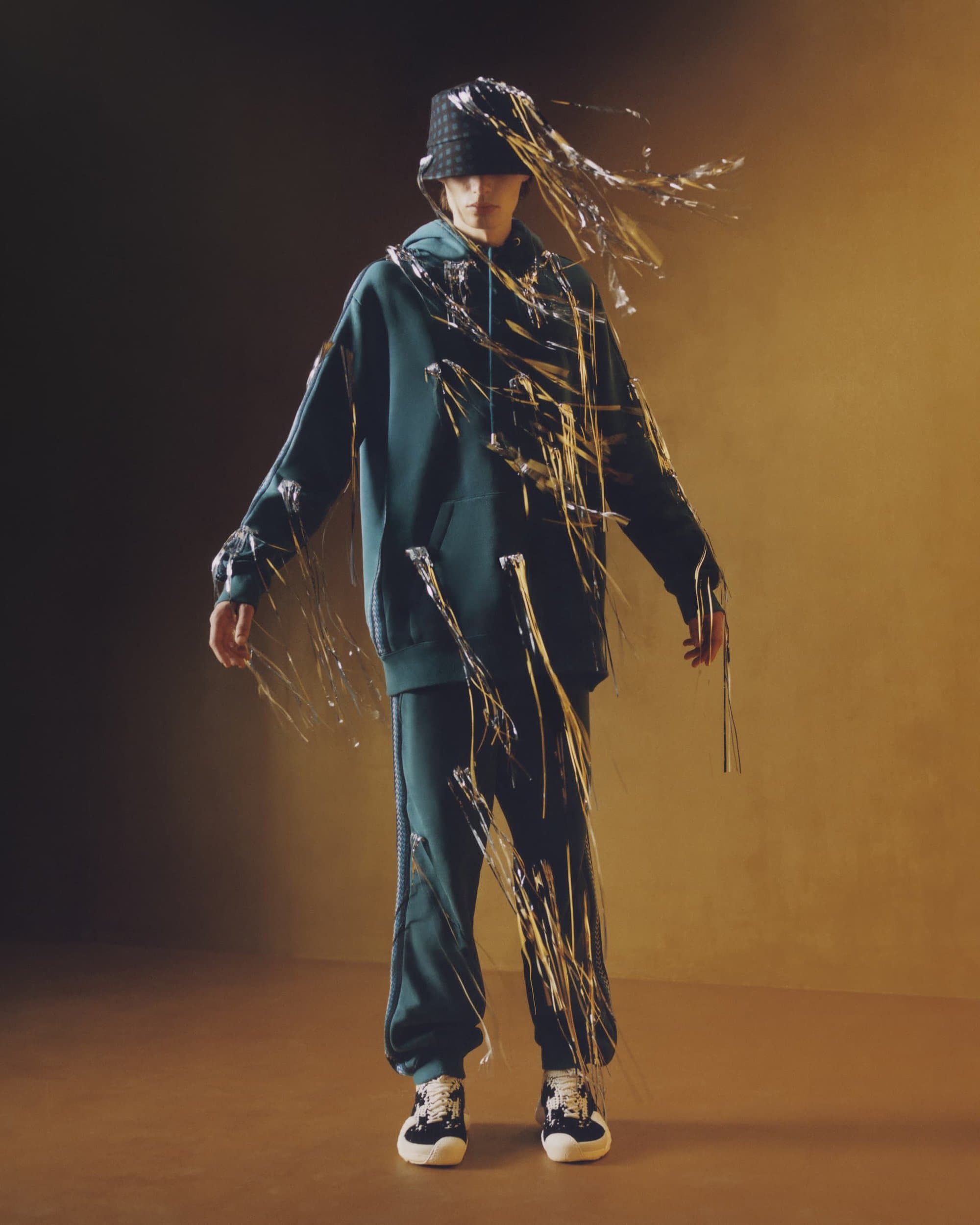 An adult poses in a studio wearing a green sweatsuit, bucket hat, and silver tinsel, standing against a gradient brown background.
