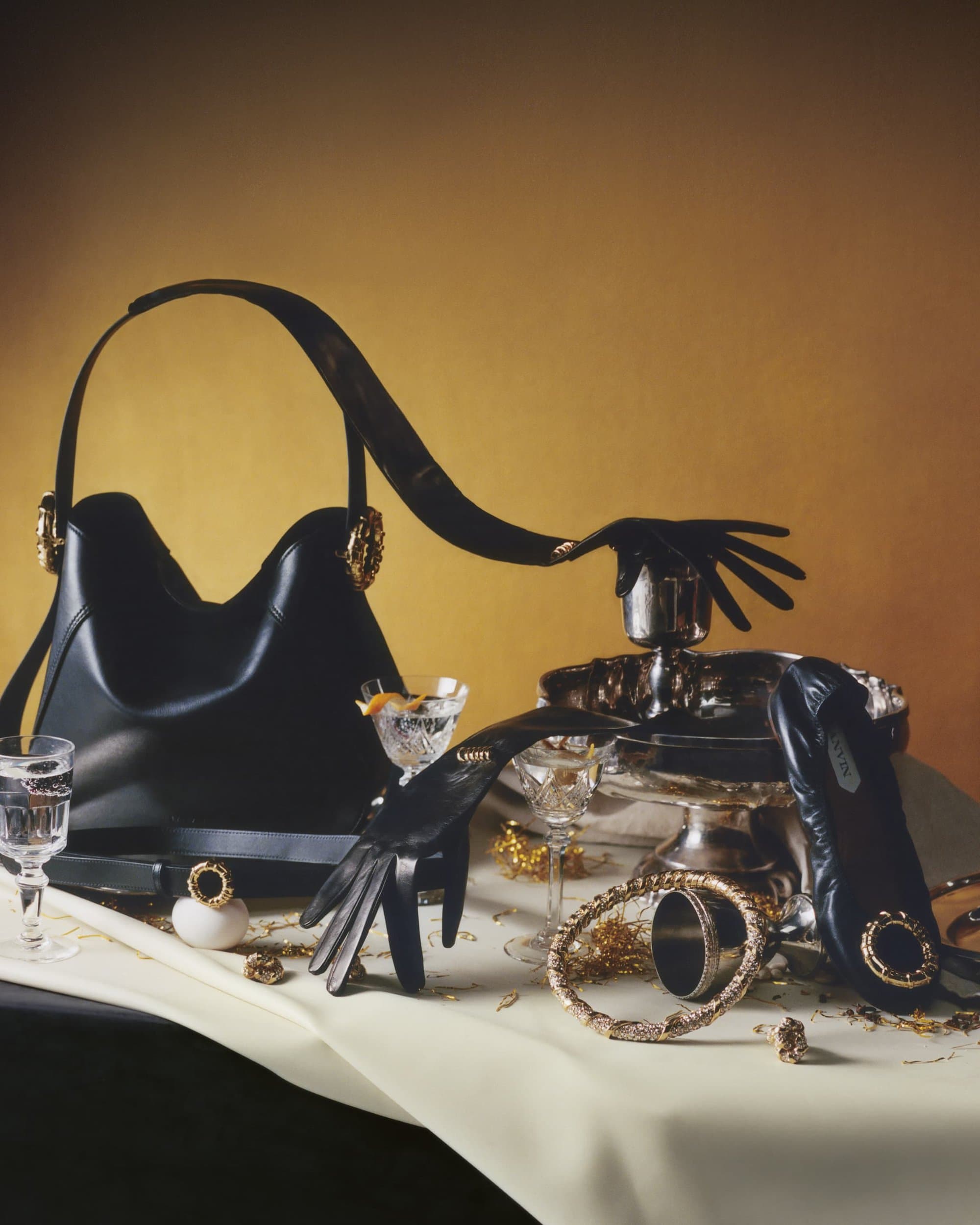 Still life composition featuring a black handbag, black leather gloves, bracelets, glassware, and other accessories on a cream-colored tablecloth.