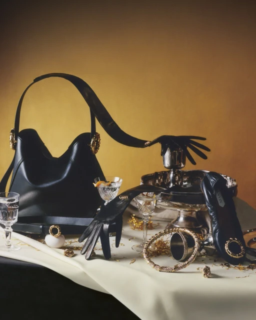 Still life composition featuring a black handbag, black leather gloves, bracelets, glassware, and other accessories on a cream-colored tablecloth.