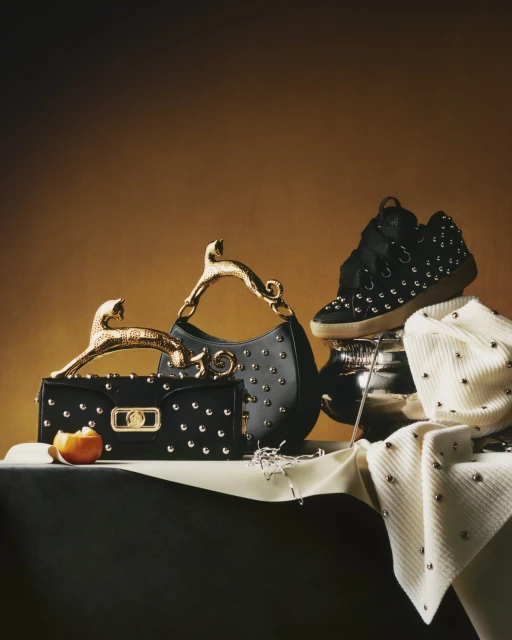 Fashion still life: studded black handbags, a studded black sneaker on a silver bowl, and a studded white sweater on a draped table.