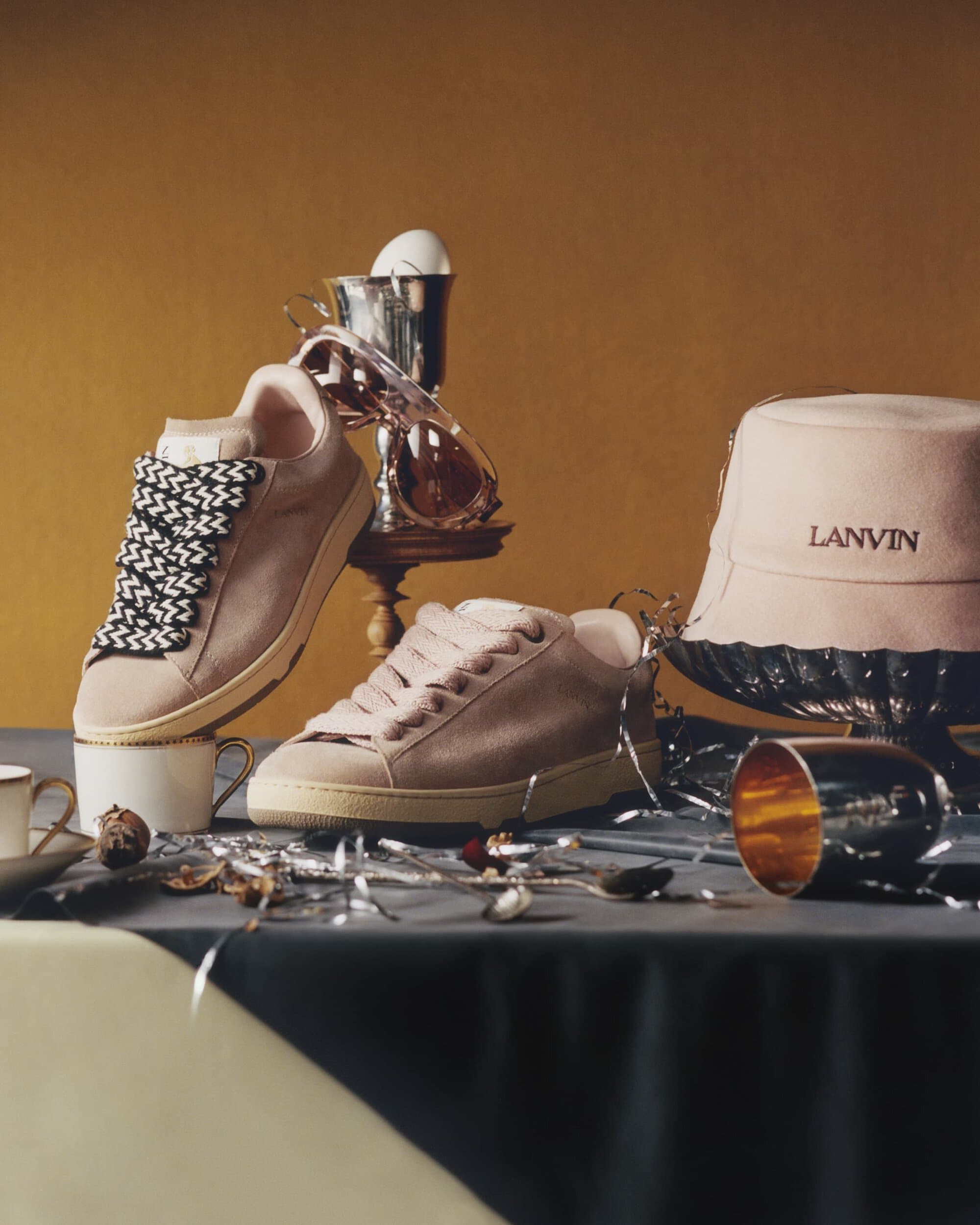 Still life composition featuring cream-colored Lanvin sneakers, sunglasses, a hat, and decorative silver objects on a draped table.