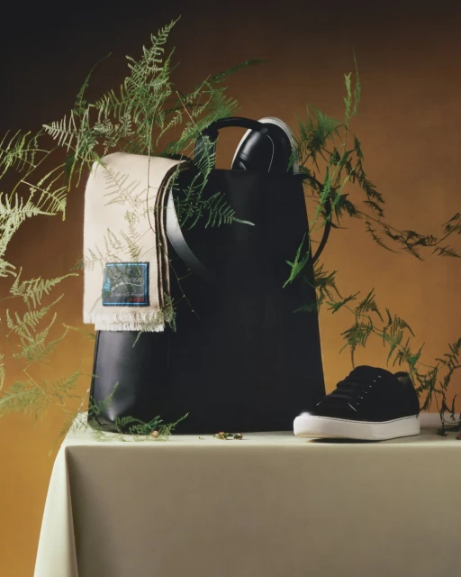 Black leather tote bag displayed with sneakers, green ferns, and a cream-colored fringed scarf on a tan draped table against an umber background.