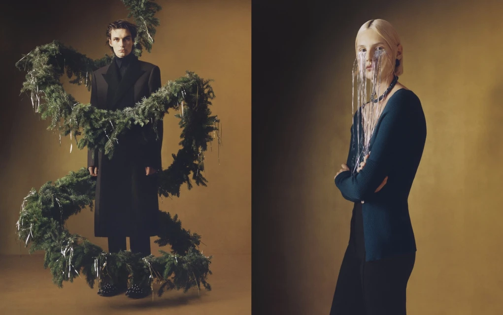 Two fashion models pose against a golden backdrop. One wears a black coat and is surrounded by a Christmas wreath; the other, a dark top and tinsel.