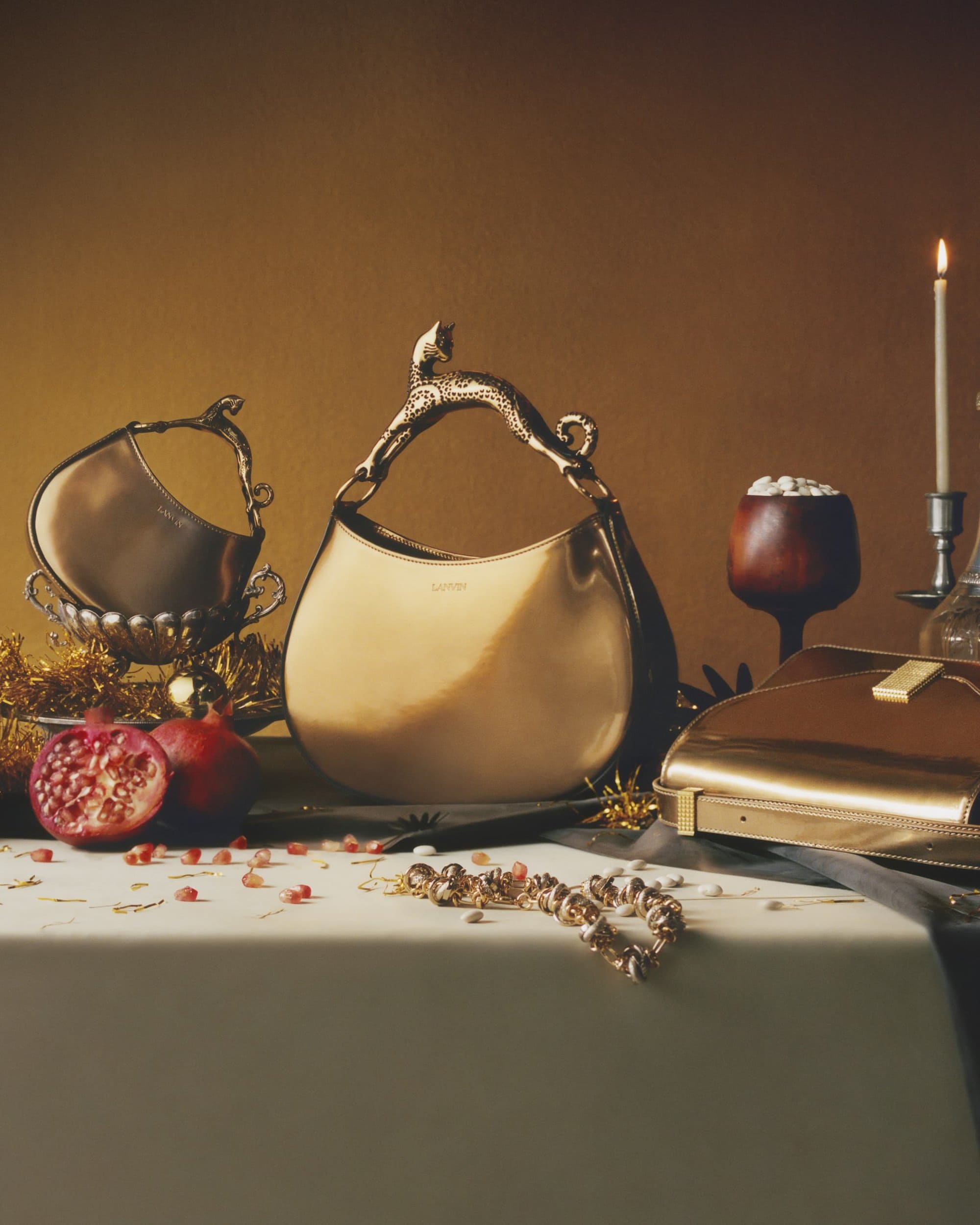 Golden handbags on a table with pomegranates, jewelry, and lit candle against a warm backdrop create a luxurious fashion still life.
