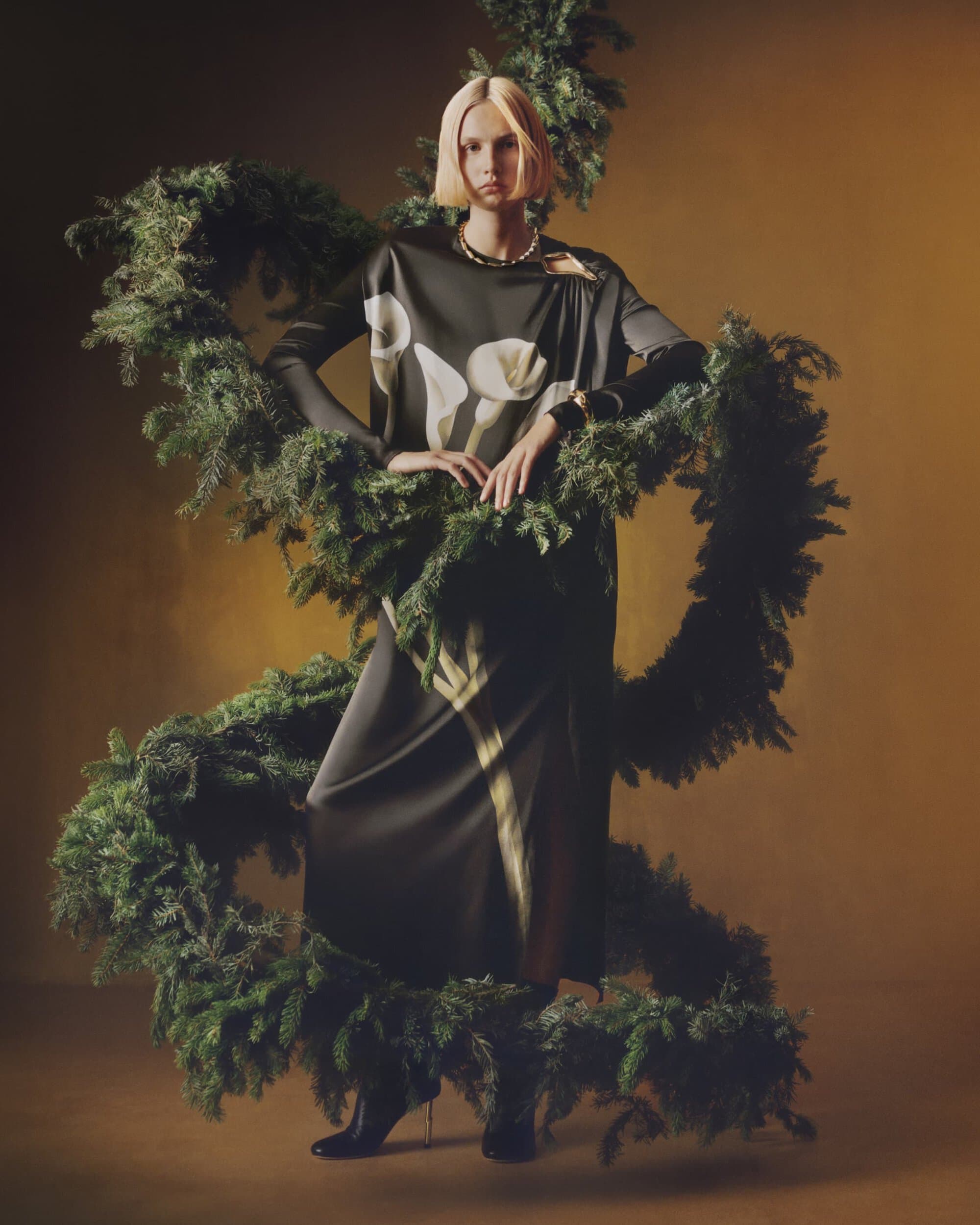 Fashion shot of a fair-skinned adult woman with short blonde hair wearing a black dress with white floral print, surrounded by green garland.
