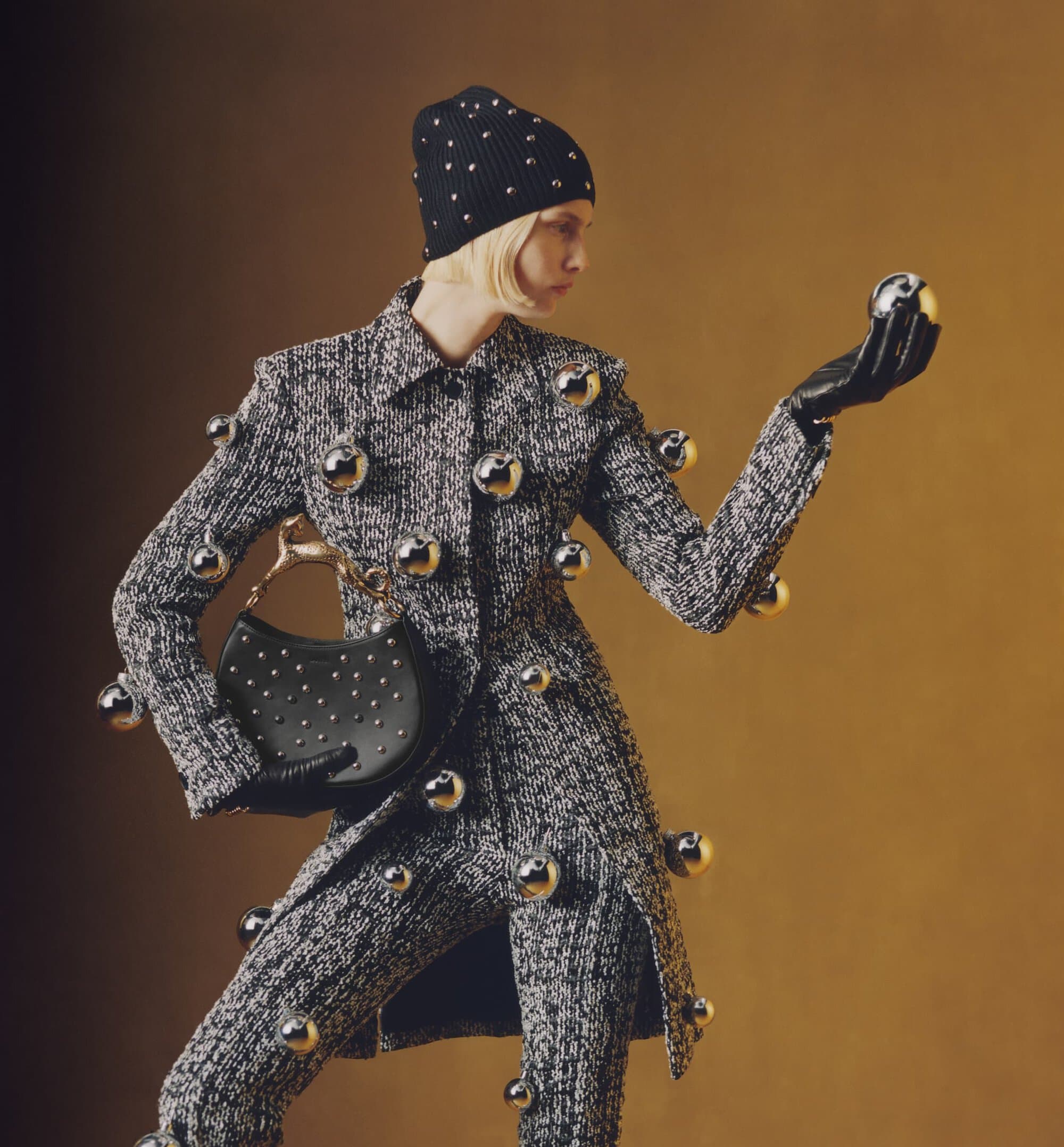 Fashion model in embellished tweed suit and beanie holds a metallic sphere, accessorized with a studded purse, set against a gradient backdrop.