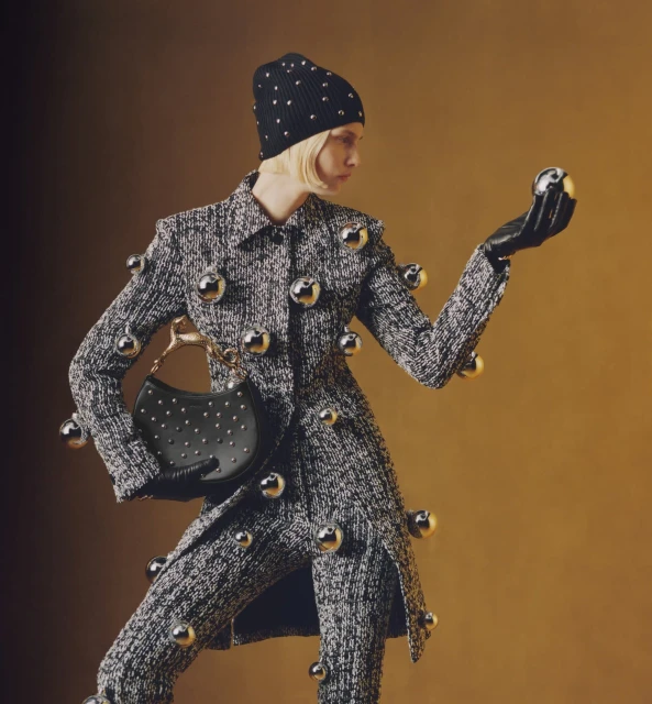 Fashion model in embellished tweed suit and beanie holds a metallic sphere, accessorized with a studded purse, set against a gradient backdrop.