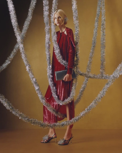 A stylish adult female stands in a studio wearing a red dress and black high heels, posing amidst silver tinsel in a fashion shot.