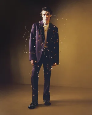 Model in a dark purple suit wrapped in fairy lights standing against a warm toned studio backdrop for Lanvin.