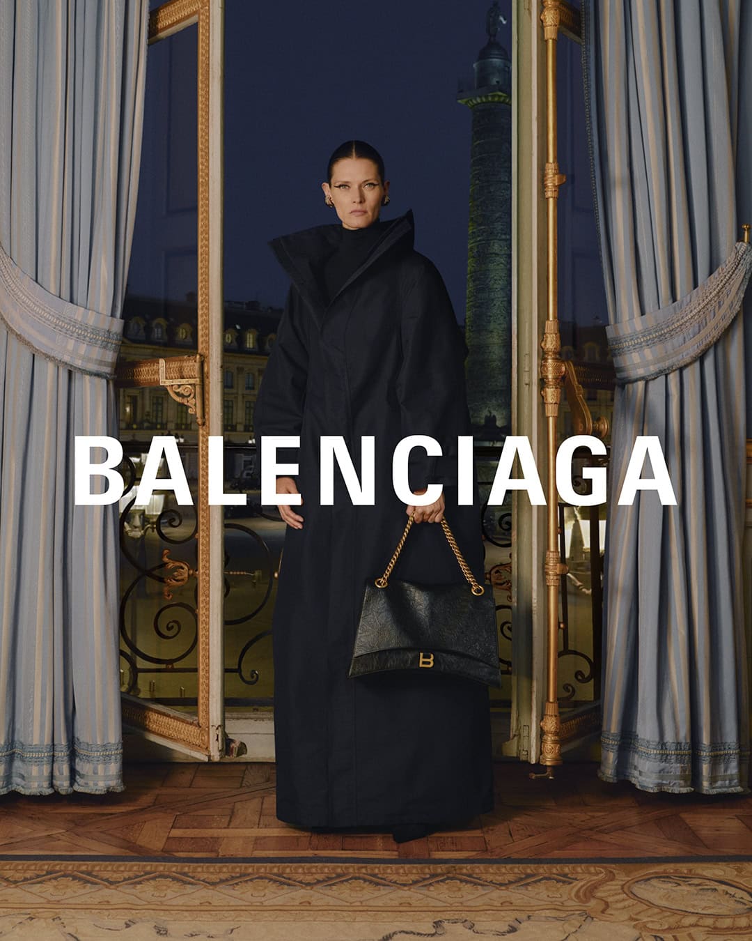 Elegant studio shot of a woman in a black coat holding a purse, standing near an ornate window with drapes, with the Balenciaga logo.