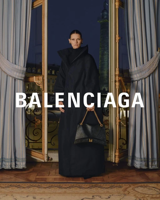 Elegant studio shot of a woman in a black coat holding a purse, standing near an ornate window with drapes, with the Balenciaga logo.