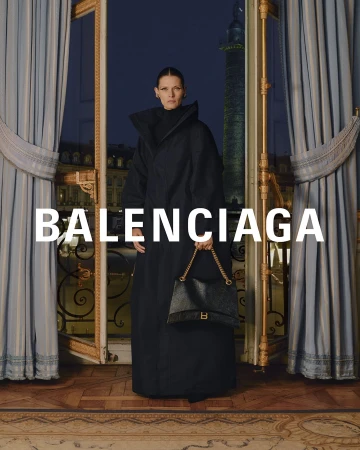 Elegant studio shot of a woman in a black coat holding a purse, standing near an ornate window with drapes, with the Balenciaga logo.
