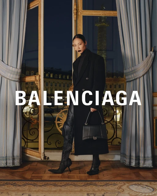 Fashion portrait of a woman in a black coat, leather boots, and handbag, posing indoors against a window overlooking a cityscape.