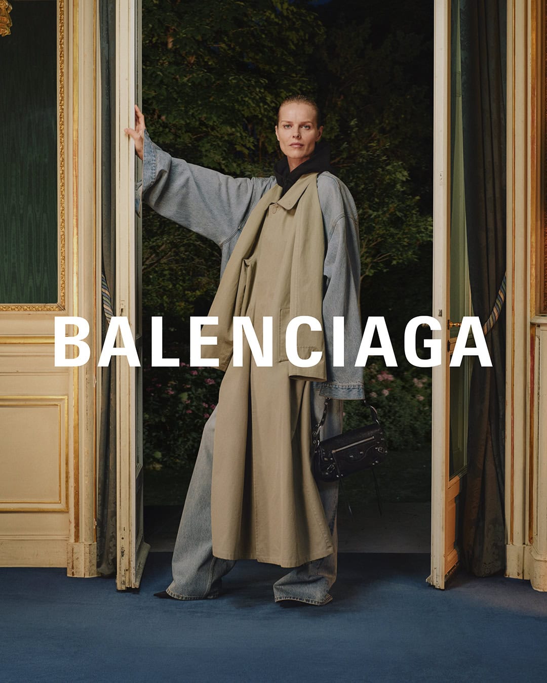 Fashion editorial shot of a woman wearing a beige trench coat, wide denim pants, and carrying a black purse, standing in a doorway.