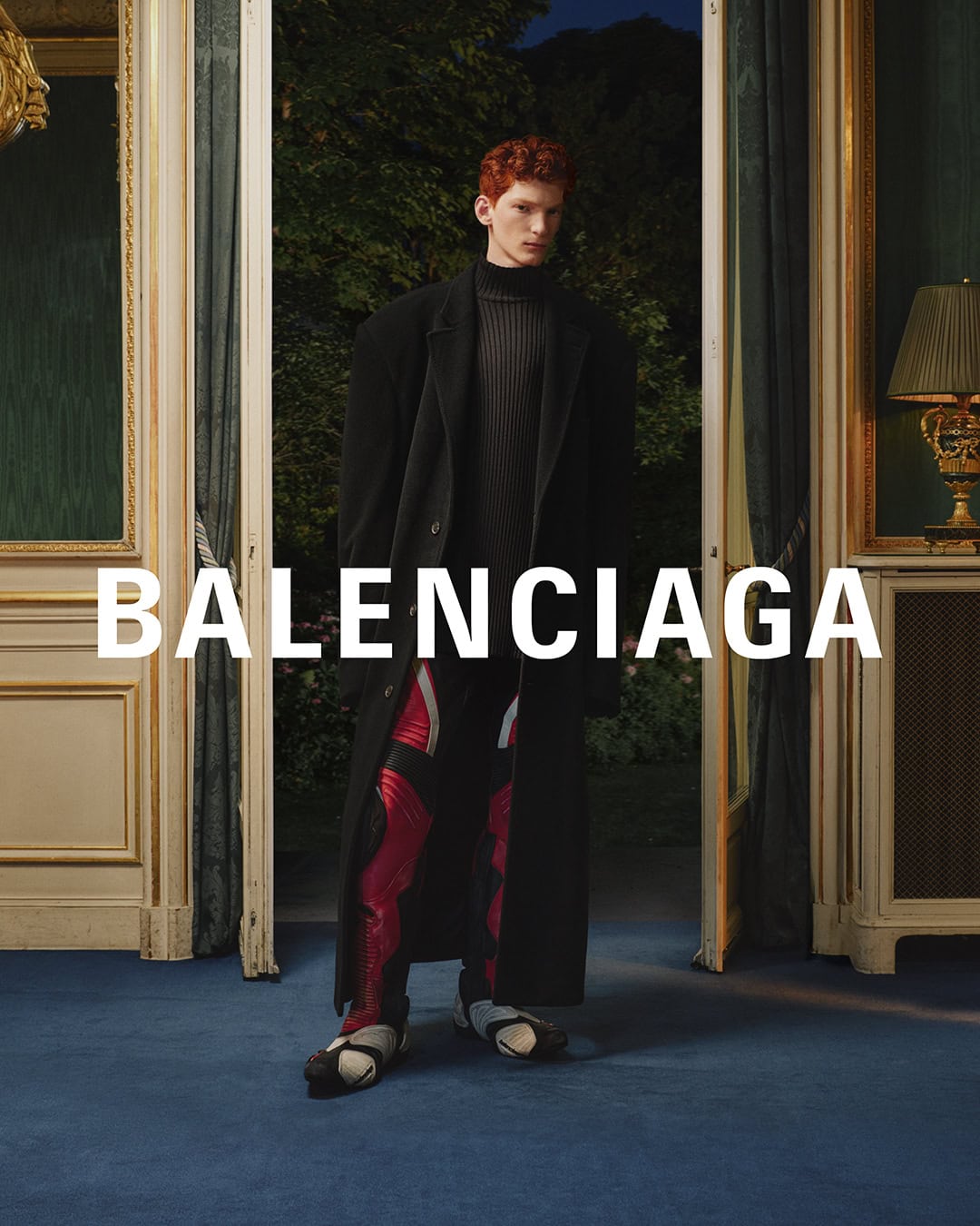 An adult male model poses in an ornate interior wearing a black coat, gray sweater, shiny red pants, and statement shoes for Balenciaga.