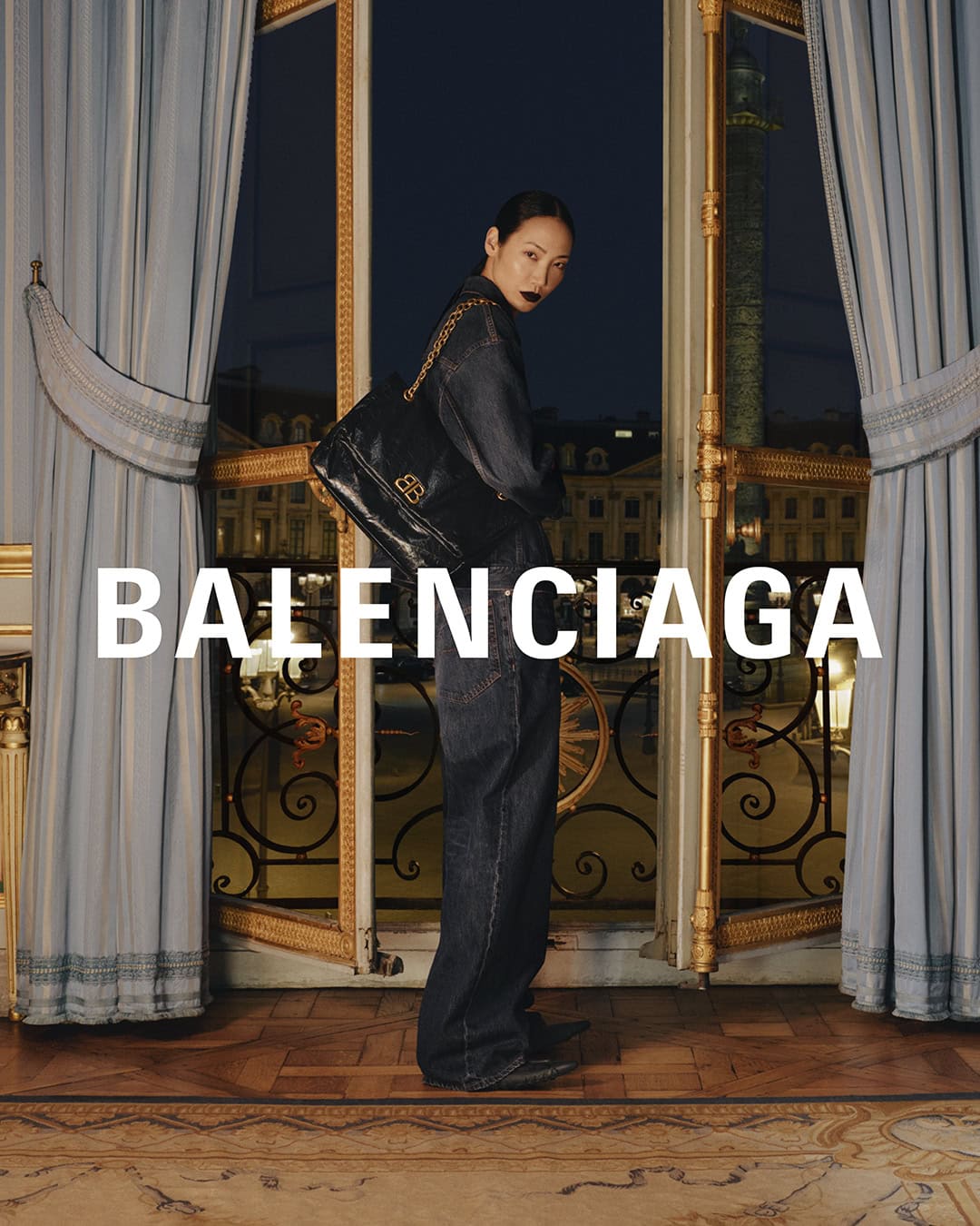 Balenciaga ad: Stylish Asian woman in denim suit with black shoulder bag, stands in ornate room with city views. Evening light.