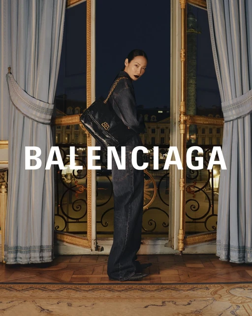 Balenciaga ad: Stylish Asian woman in denim suit with black shoulder bag, stands in ornate room with city views. Evening light.