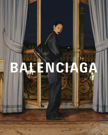Balenciaga ad: Stylish Asian woman in denim suit with black shoulder bag, stands in ornate room with city views. Evening light.