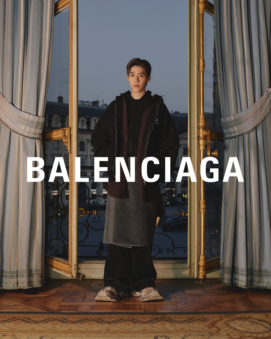 Asian male fashion model wears Balenciaga in an indoor scene with natural lighting, standing near a window overlooking a cityscape.