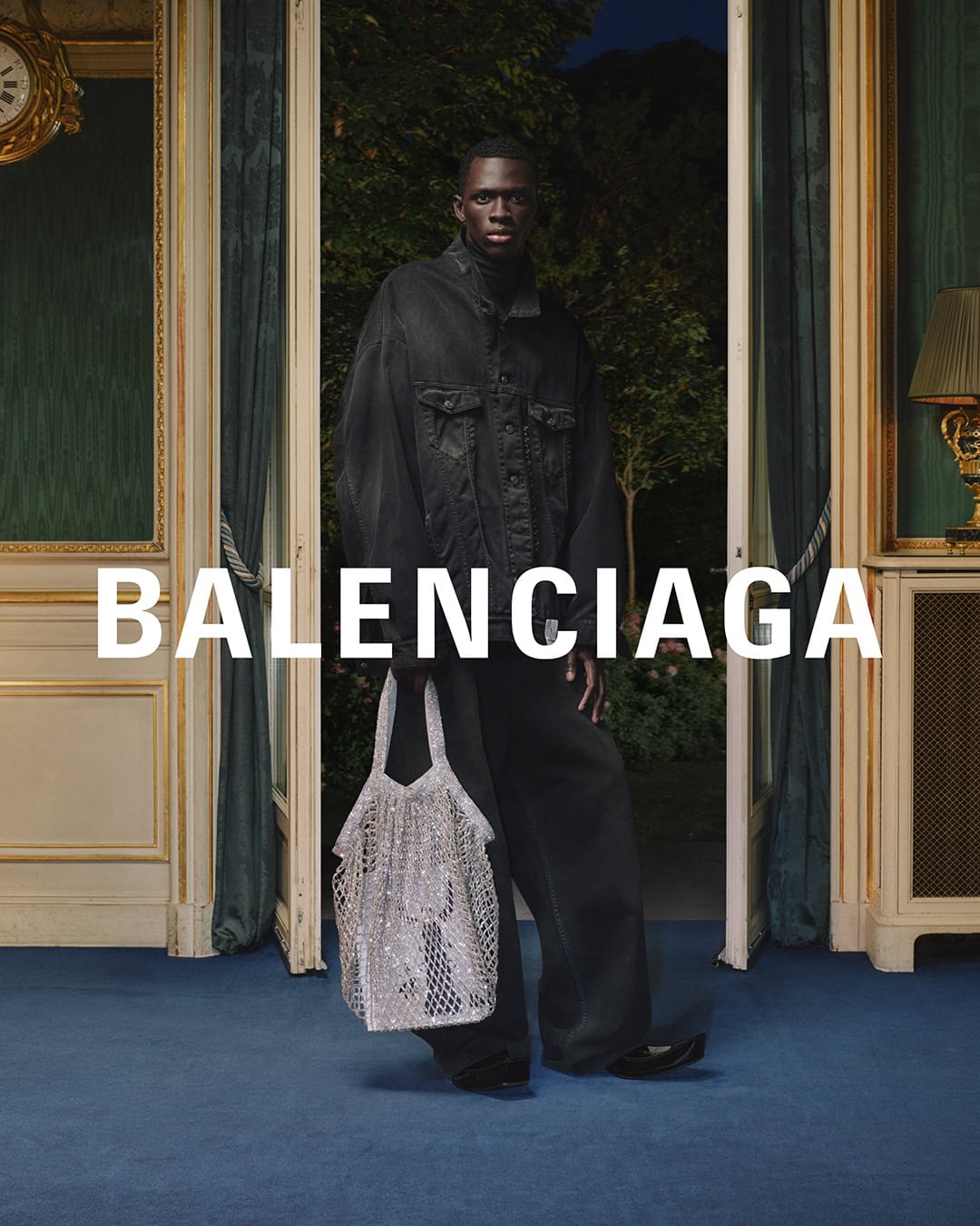 An adult male poses indoors wearing a dark jacket and pants while holding a silver mesh bag, with a large BALENCIAGA text overlay.