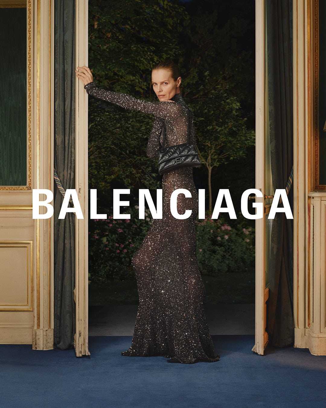 Woman poses in glittering gown and small quilted purse, standing in doorway with blue carpet, advertising the fashion brand Balenciaga.