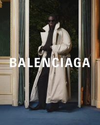 Model wearing dark sunglasses and an oversized white trench coat standing in a doorway for Balenciaga Spring 2024 campaign