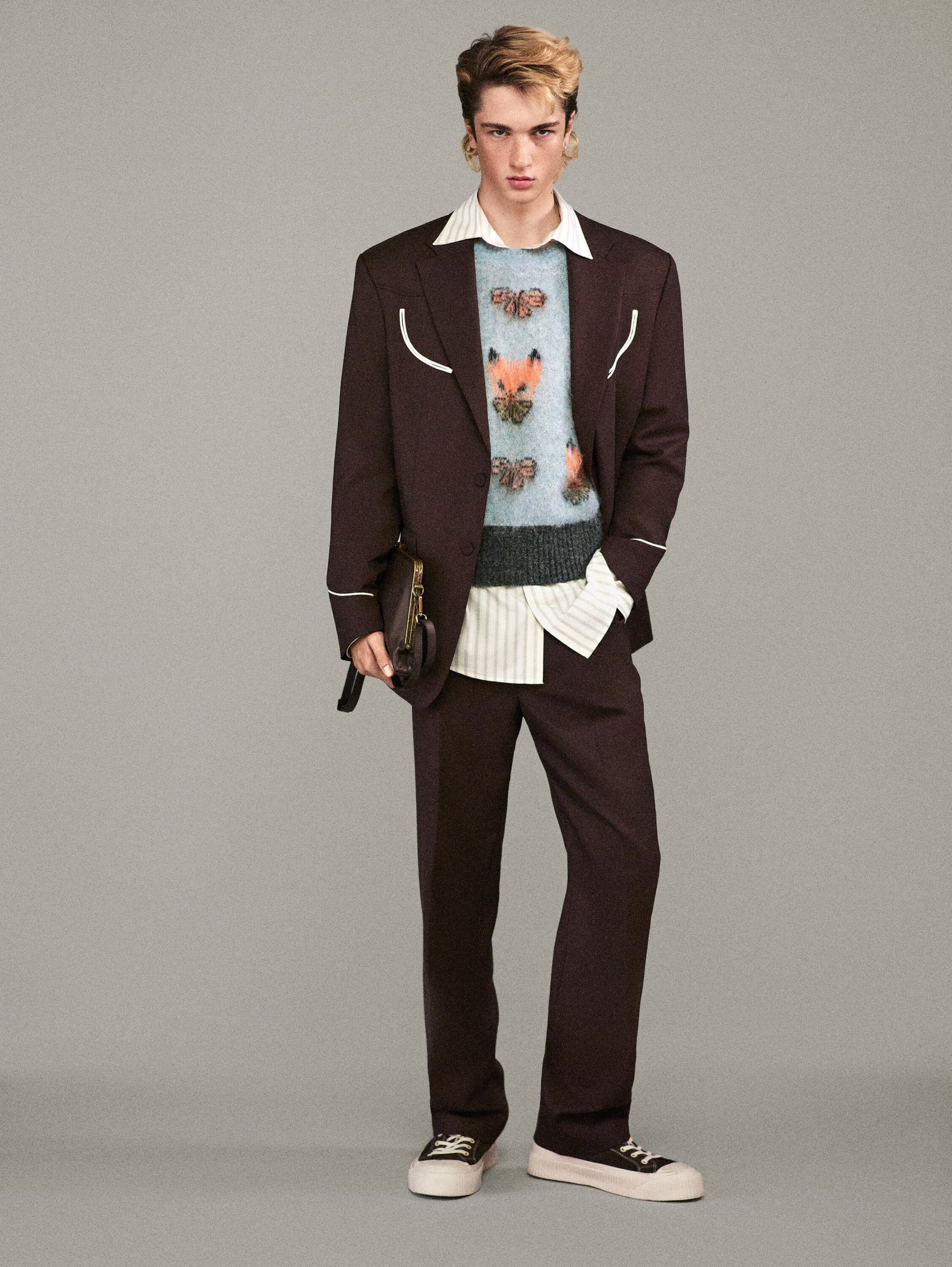 Full-length shot of a young adult man in a brown suit with a sweater with animal designs, posing in front of a solid grey background.