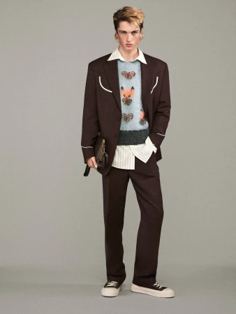 Full-length shot of a young adult man in a brown suit with a sweater with animal designs, posing in front of a solid grey background.
