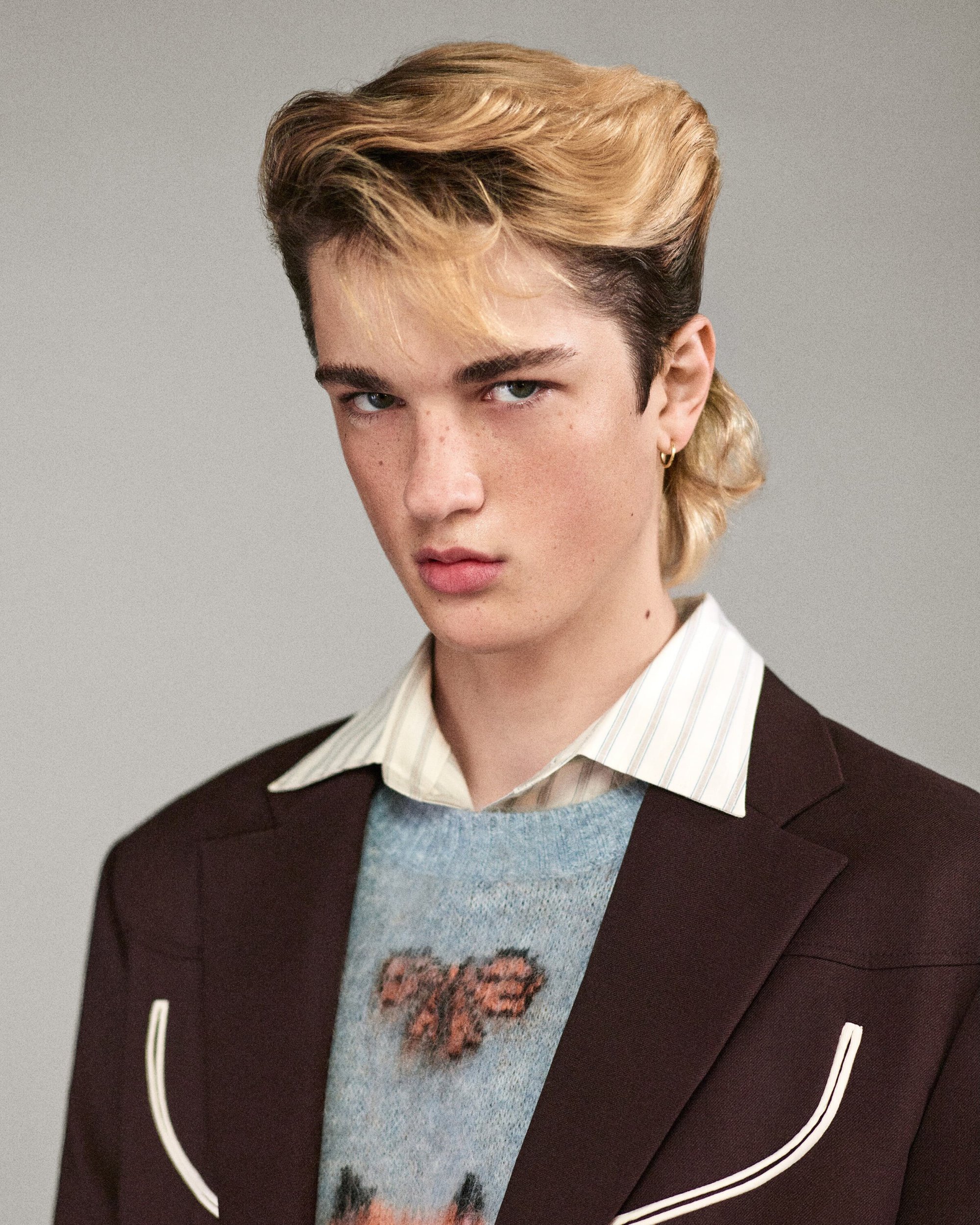 Young man with mullet hairstyle in a brown blazer and patterned sweater poses indoors against a neutral background in a portrait shot.