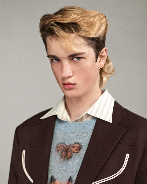 Young man with mullet hairstyle in a brown blazer and patterned sweater poses indoors against a neutral background in a portrait shot.