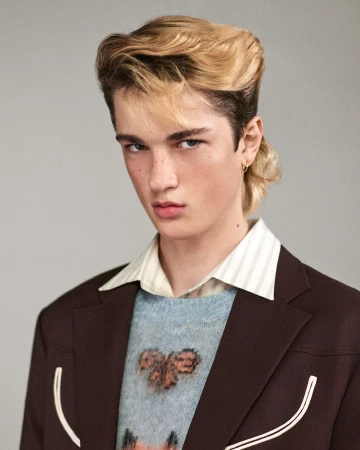 Young man with mullet hairstyle in a brown blazer and patterned sweater poses indoors against a neutral background in a portrait shot.