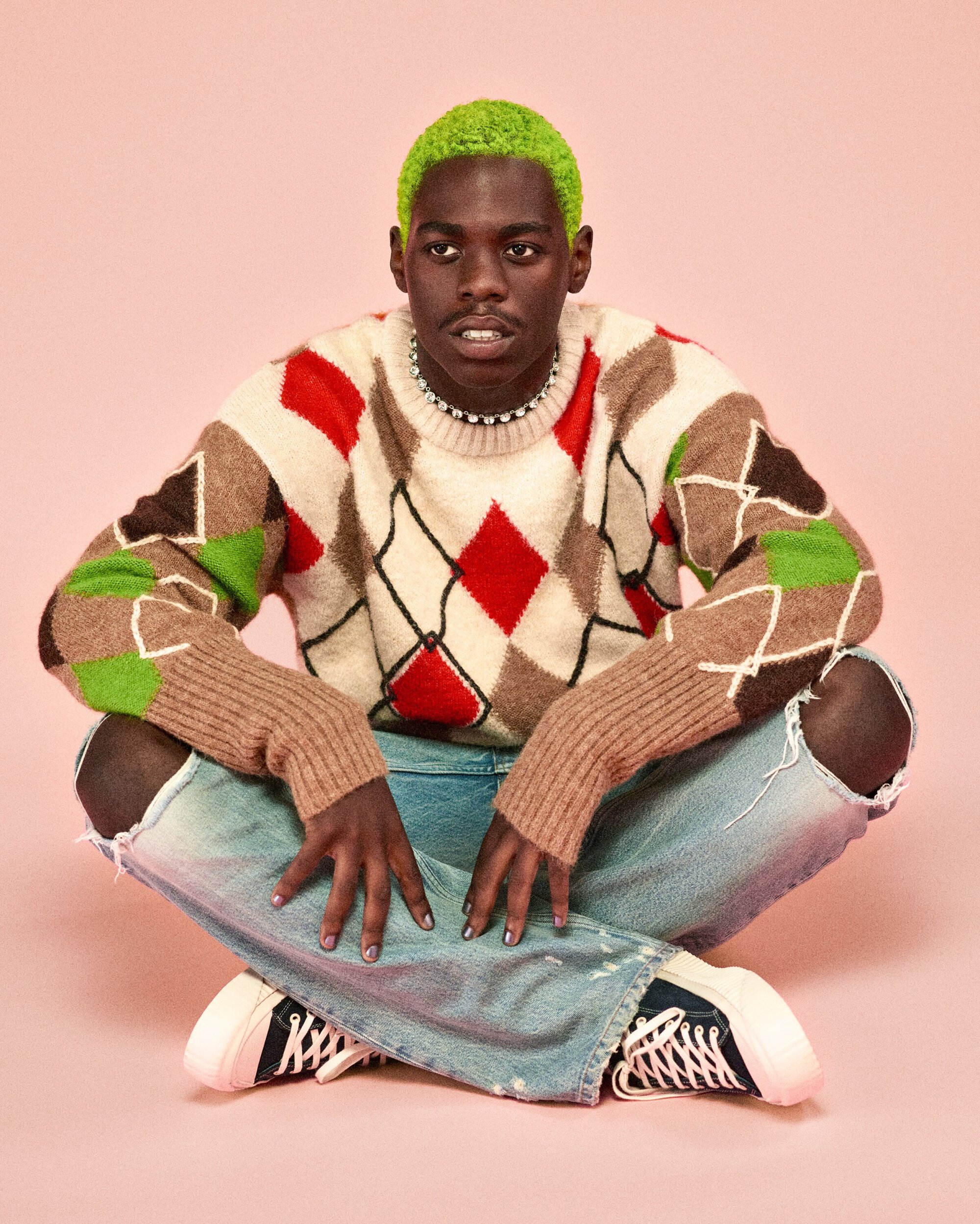 Portrait of a young adult with green hair, wearing an argyle sweater and ripped jeans, sitting cross-legged against a pink background.