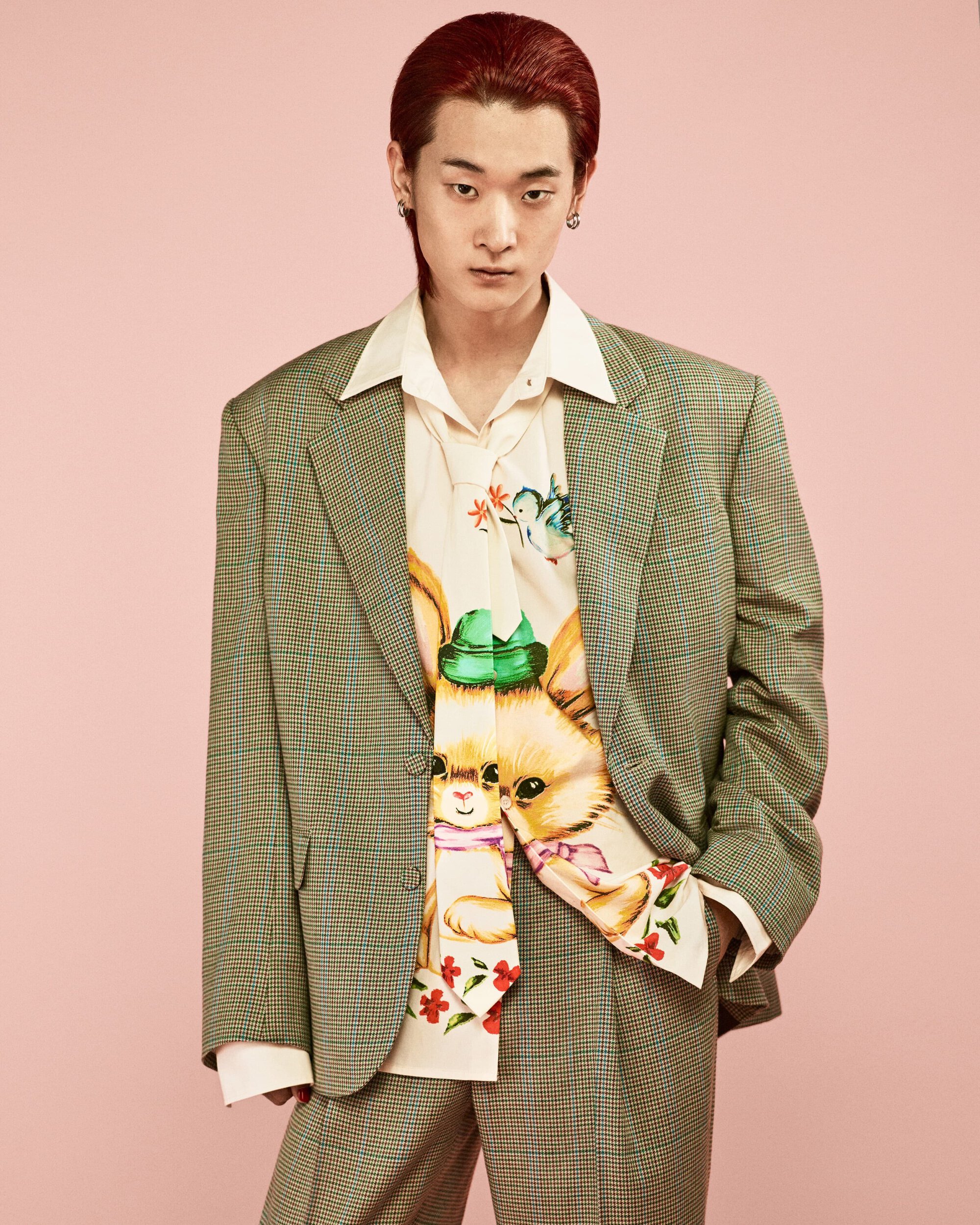 Man in an olive green suit and bunny-print shirt poses in front of a pink background in a portrait-style studio fashion shoot.