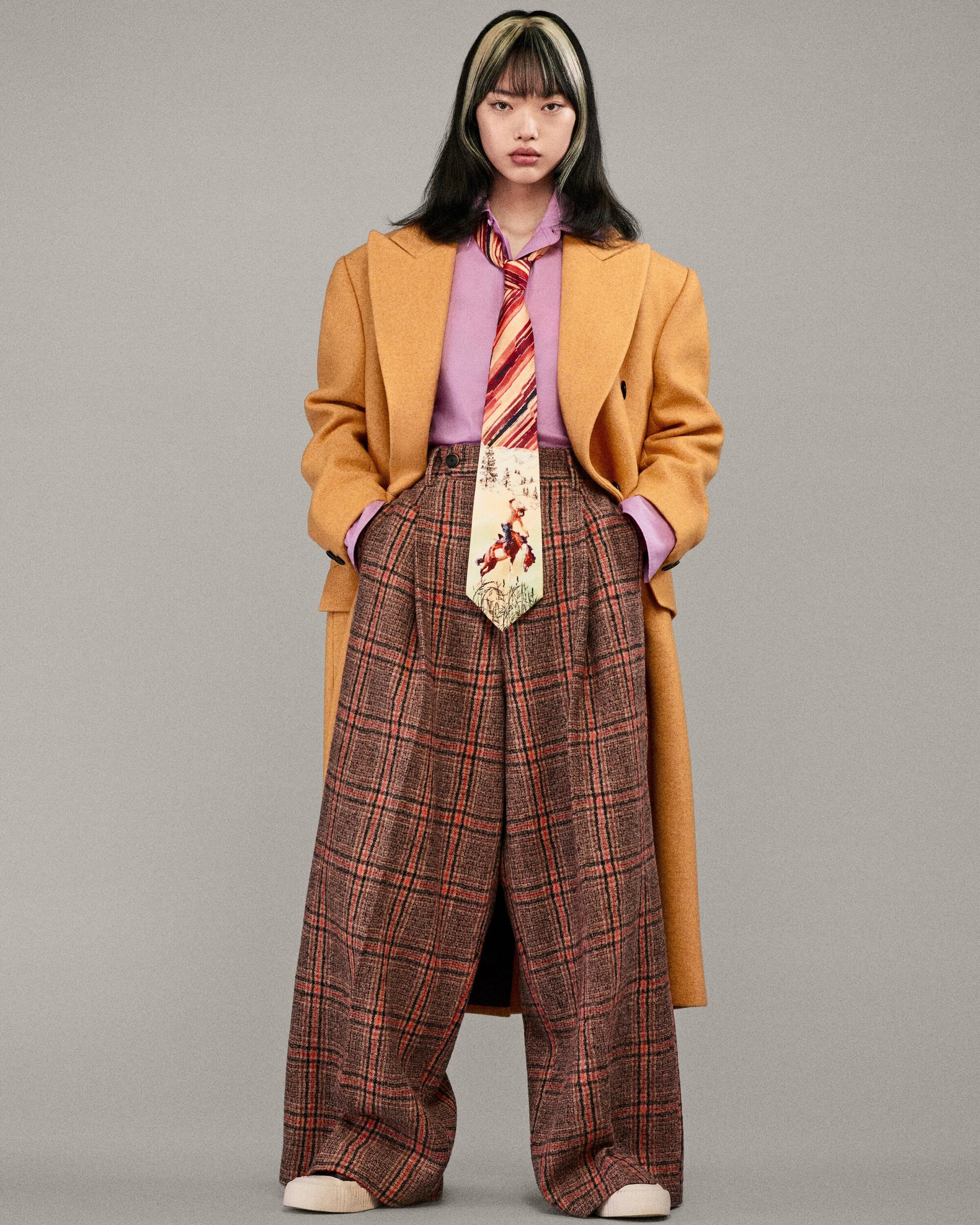 Young woman in oversized yellow coat, purple shirt, patterned tie, and plaid pants, posing in a studio against a neutral gray backdrop.