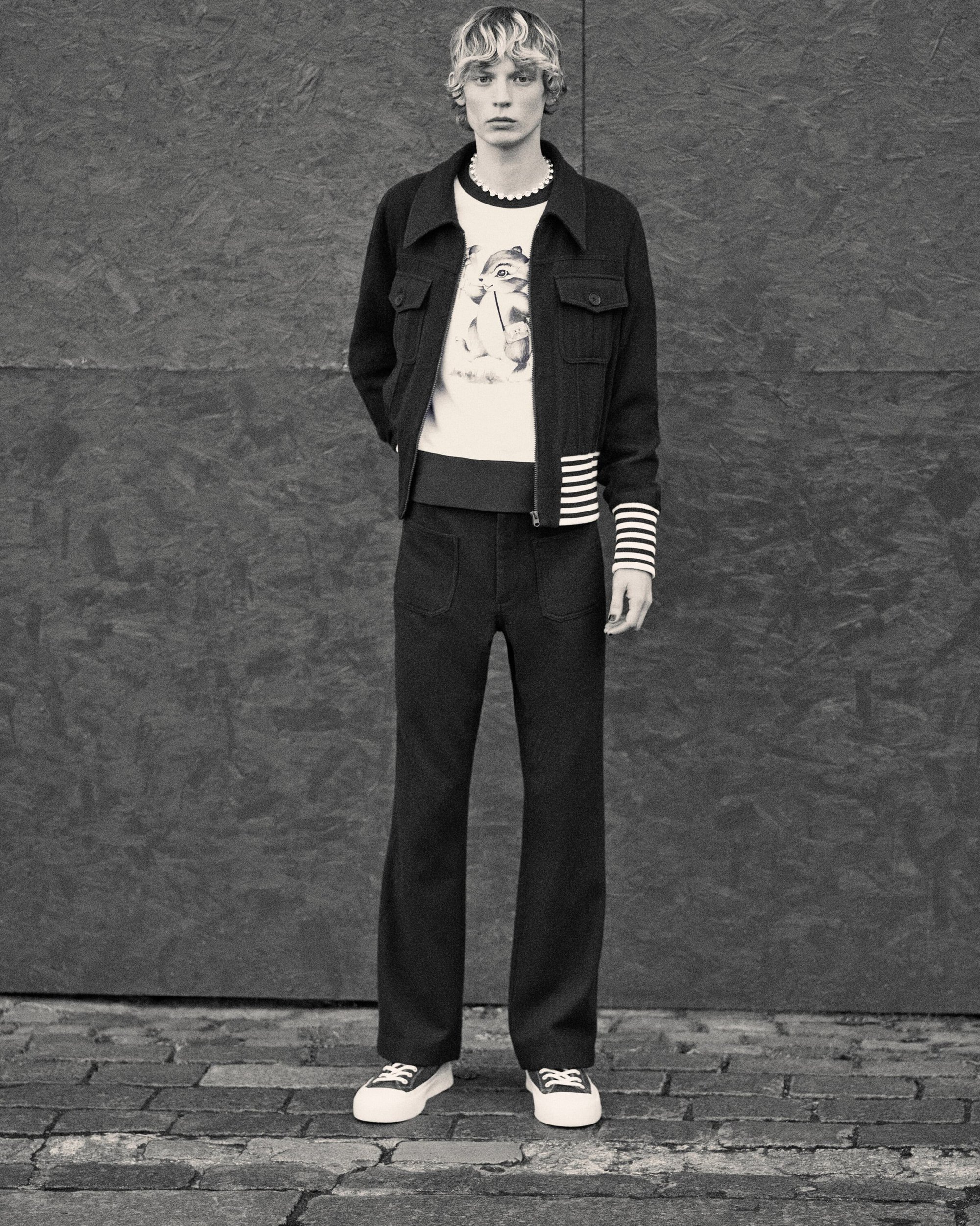 Full-length studio shot of a fair-skinned male adult model wearing dark jacket, cream sweater, dark pants, and sneakers against a grey wall.