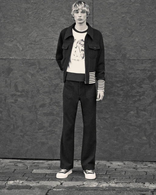 Full-length studio shot of a fair-skinned male adult model wearing dark jacket, cream sweater, dark pants, and sneakers against a grey wall.